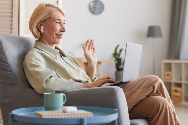 Online communication at home Mature woman sitting on armchair with laptop looking at computer monitor and waving to her friend