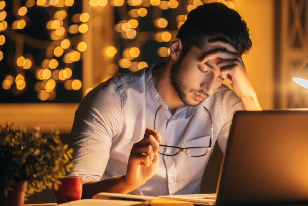 Feeling exhausted. Frustrated young man carrying eyeglasses and keeping eyes closed while sitting at his working place at night time with Christmas lights in the background Frustrated young man carrying eyeglasses and keeping eyes closed while sitting at his working place at night time with Christmas lights in the background