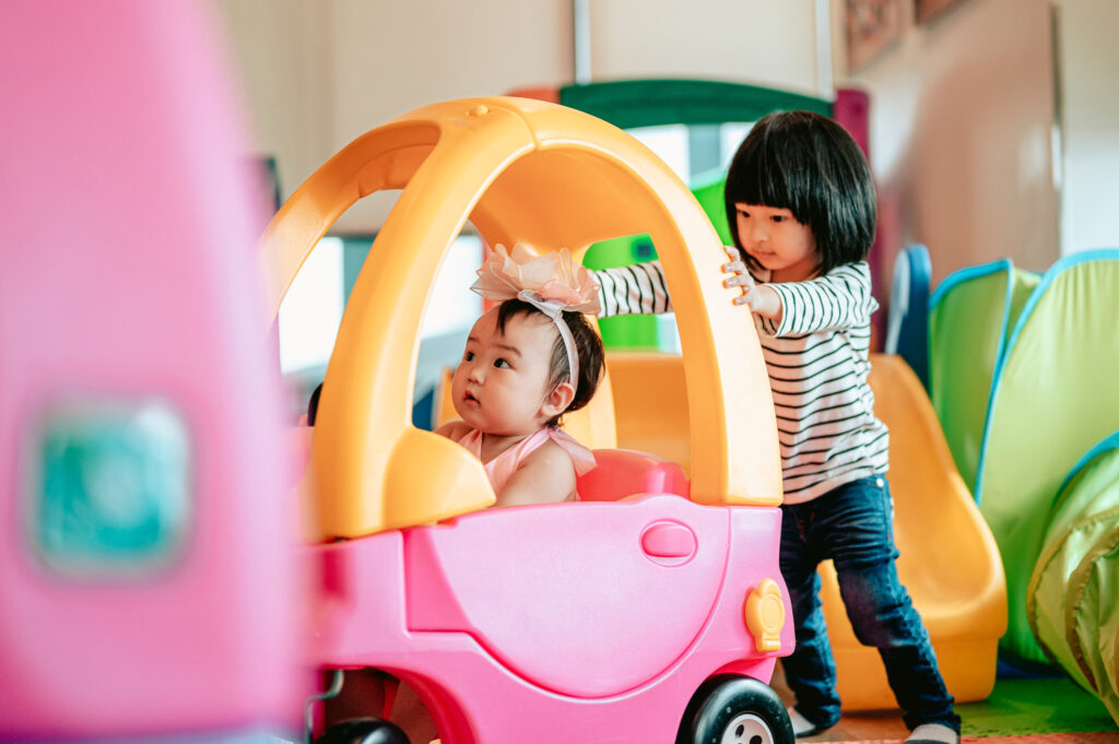 Two toddlers playing with one sitting in a pink toy vehicle and the other pushing. 