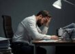 Man at desk, with head in palm, looking overwhelmed by stacks of paperwork.